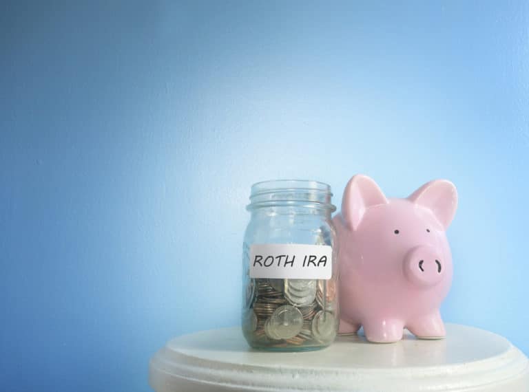 Jar containing money labeled Roth IRA sitting on a table next to a piggy bank in front of a blue background